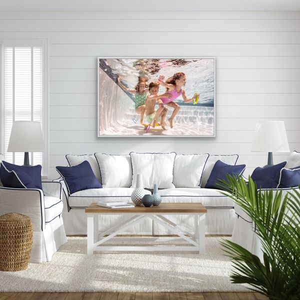 Framed acrylic print of three children playing underwater, hung above a white sofa with navy pillows, DIVE IN artwork by Leigh Webber