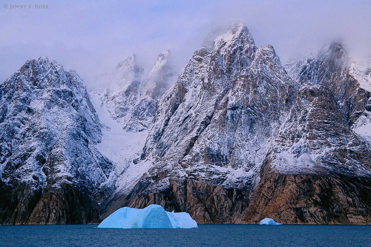 Fog-shrouded mountain spires dusted with snow soar above melting icebergs in Ofjord within Scoresby Sund, East Greenland.