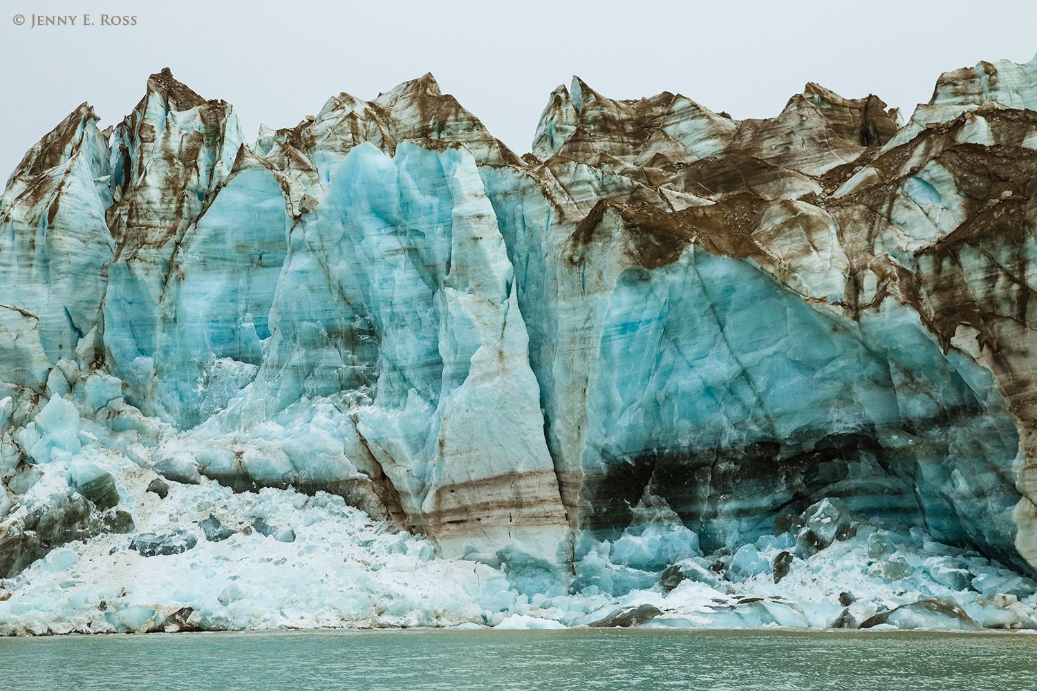 As the ice of Sefstrom Glacier disintegrates and recedes, its surface is increasingly coated with residual dirt and rock debris. Clean blue ice is exposed where icebergs have broken off the glacier face.