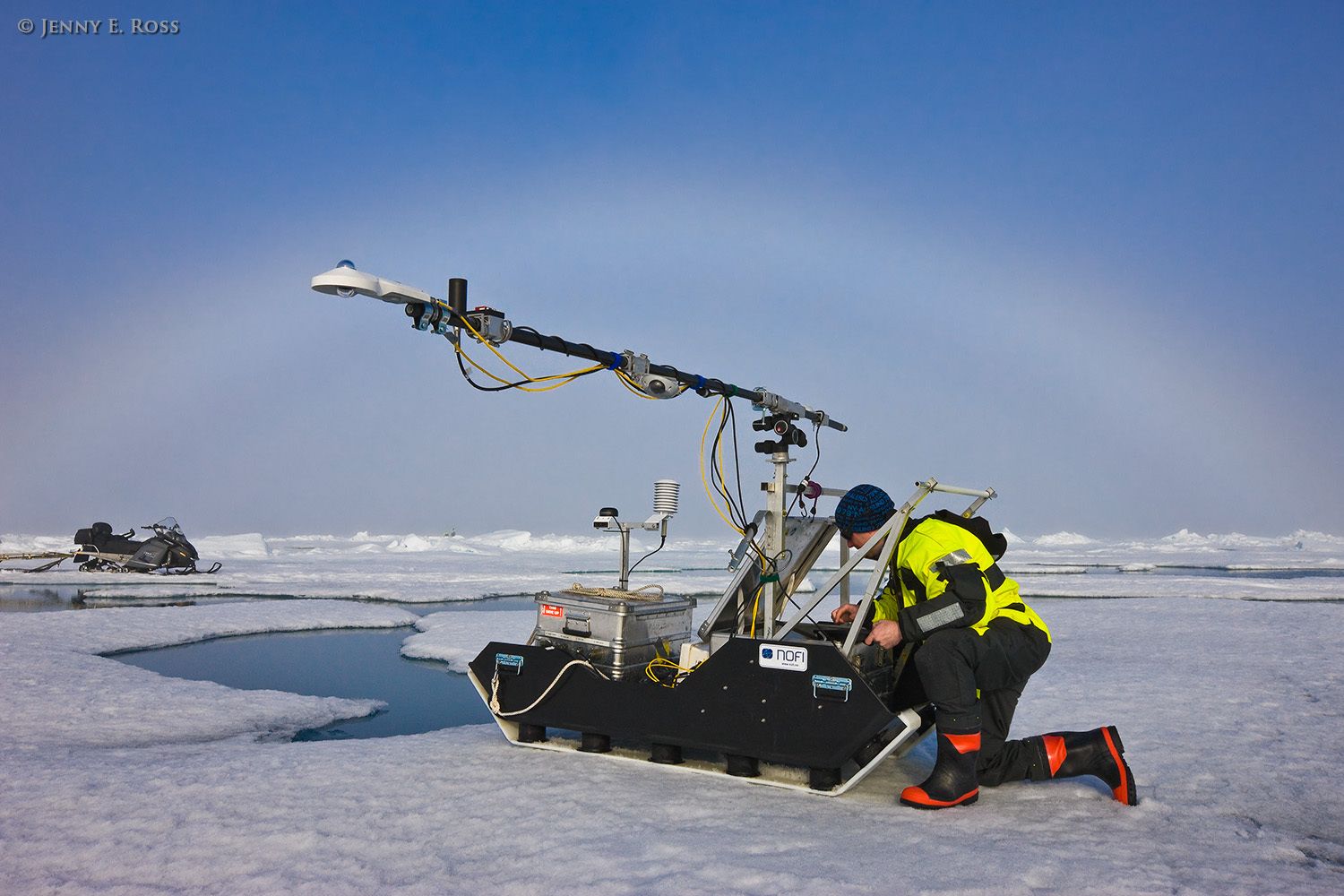 Norwegian Polar Institute geophysicist Dr. Stephen Hudson sets up several scientific devices on a portable sled to make measurements relating to the absorption and reflection of solar energy by various forms of Arctic sea ice. The scientist and his colleagues collected data on a large floe of melting summer sea ice in the Arctic Ocean as part of NPI's 2012 "ICE" (Ice, Climate, and Ecosystems) expedition. The areas of water in the photograph are shallow melt ponds of varying depth on the surface of the ice floe. The NPI ship RV Lance was attached securely to this particular floe of ice, and the vessel moved with the floe as the ice floated freely in the ocean, for the duration of various on-ice research activities in July-August 2012. A fog bow arches across the sky in the distance. Scientific research on arctic sea ice, central polar basin, Arctic Ocean