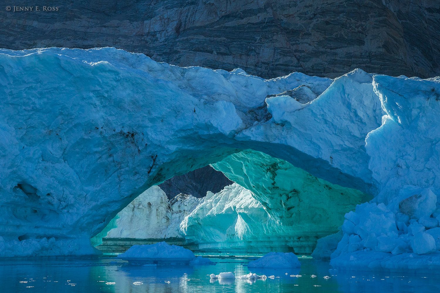 In the early morning, an arched iceberg floats beneath a towering cliff of ancient metamorphosed sedimentary rock near Daugaard-Jensen Glacier within Norvest Fjord in Scoresby Sund, Northeast Greenland National Park, Greenland.