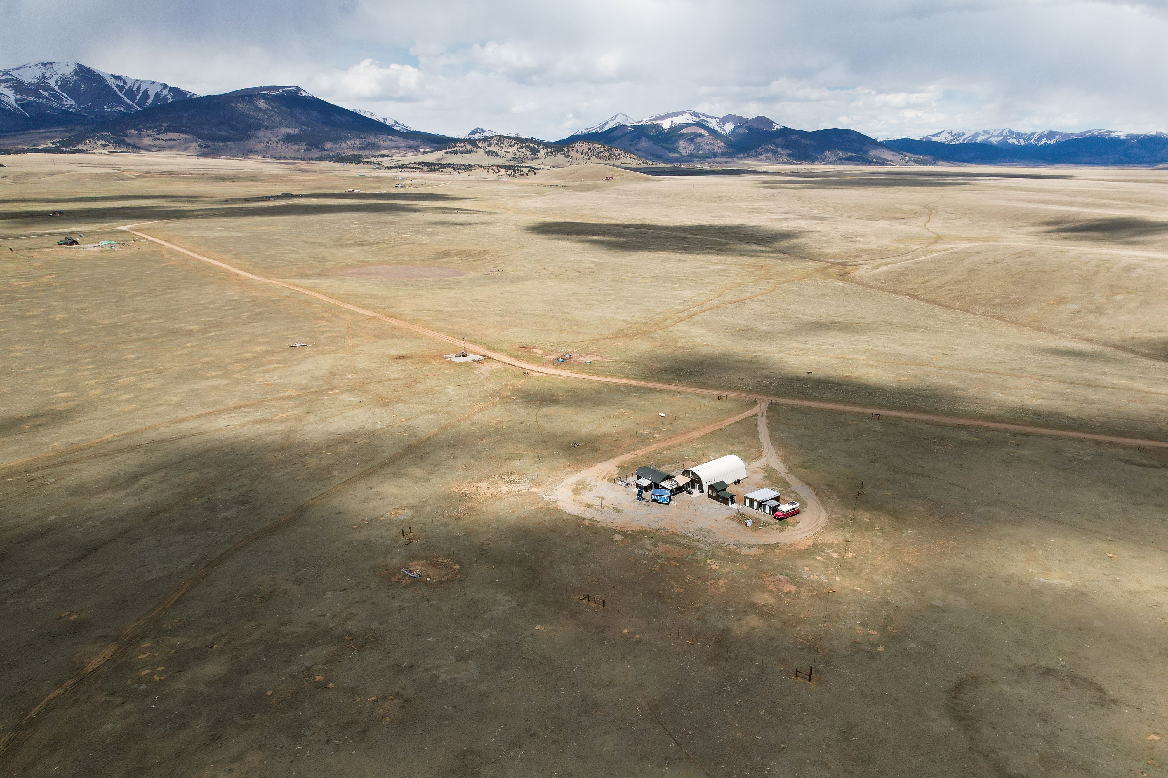 A very high view taken by a  drone of a Colorado homestead property with a heart-shaped driveway. Clouds make shadows on the surrounding landscape with snow capped Rocky Mountains on the horizon. Heart-Shaped Driveway