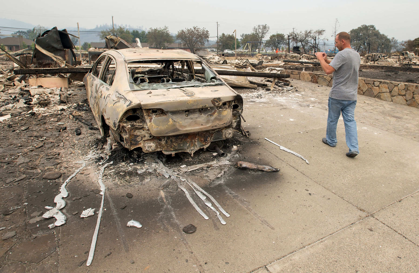 A man takes a photo of his daughter's burned out new car in her driveway after a fire tore through the area.