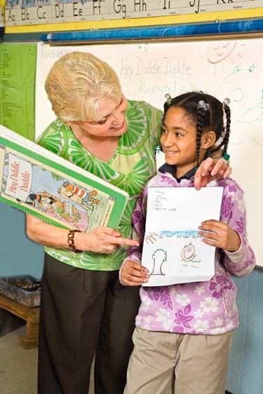 Blonde female teacher smiles and points to drawing and spelling list held by African American female second grade elementary smiling student in classroom Female teacher points to drawing held by happy black girl in class