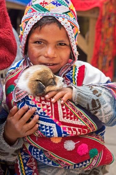 Peruvian young girl in hand embroidered costume dress and hat holds puppy close to her. Peruvian child in colorful dress holds puppy
