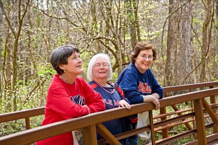 Three mature female friends wearing Ole Miss shirts rest on wooden country bridge in William Faulkner's Woods in Oxford, Mississippi Three older women lean on bridge in woods