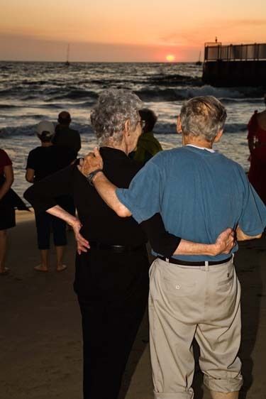 Mature man and woman with gray hair stand on beach holding arms around each other looking at sunset with other people at dusk at Playa del Rey beach in Los Angeles, California Senior couple wrap arms around each other looking at sunset on beach