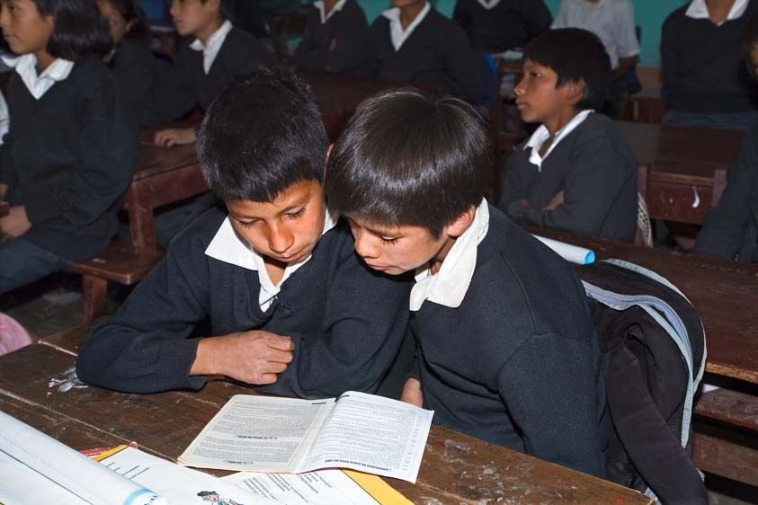 Two Peruvian elementary age boys in uniforms work together at a desk for two in a school classroom reading a book for their lesson. Two Peruvian school boys share lesson in class