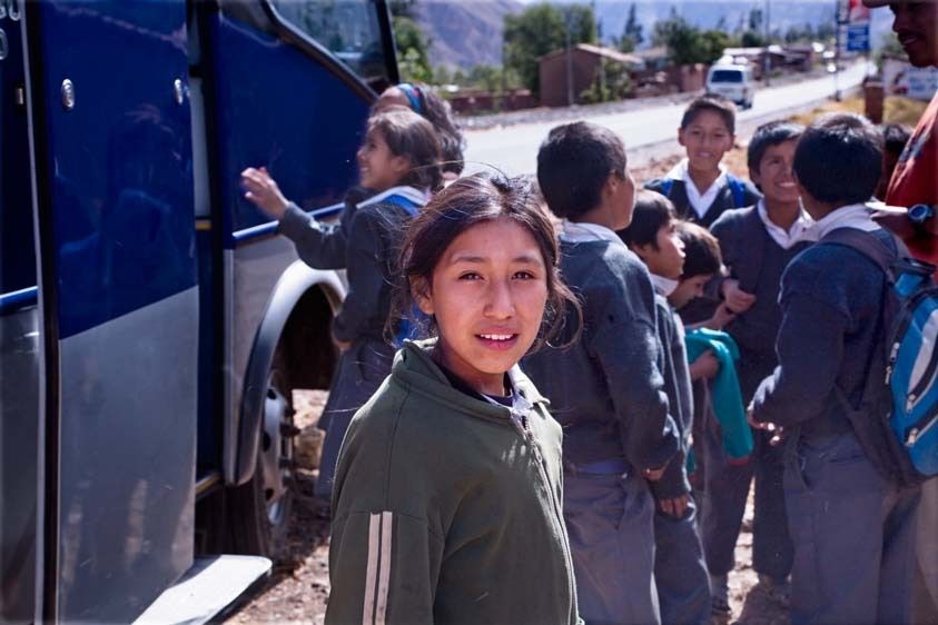 Peruvian elementary age girl in jacket waits with school boys and girls in uniforms at bus stop in Peru in mountains Peruvian female student at bus stop and school children
