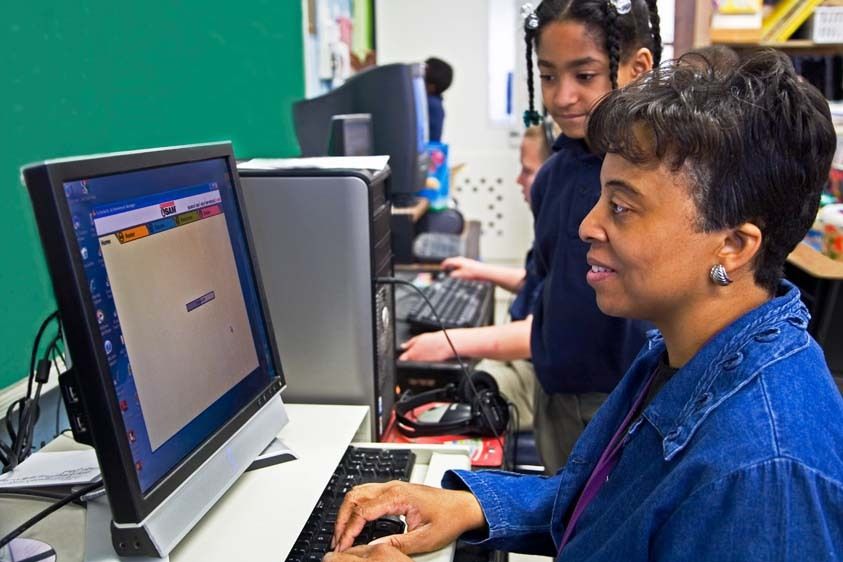 African American female teacher demonstrates to African American second grade girl program on computer in classroom in New Orleans, Louisiana. Black woman teacher instructs black female student on computer