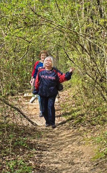 Two senior female friends wear Ole Miss shirts and walk on path through green shrubs and trees in William Faulkner Woods in Oxford, Mississippi Senior women hike through woods