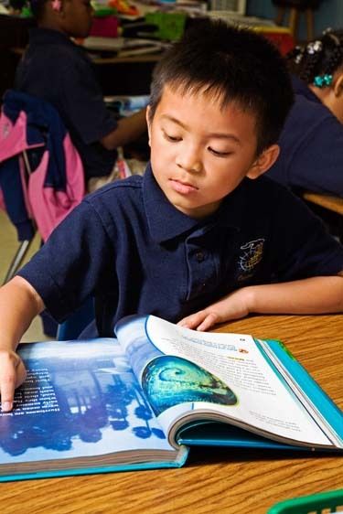 Vietnamese American second grade male sits at desk in classroom and points to word on page in book in New Orleans, Louisiana Vietnamese boy seven years old reads book