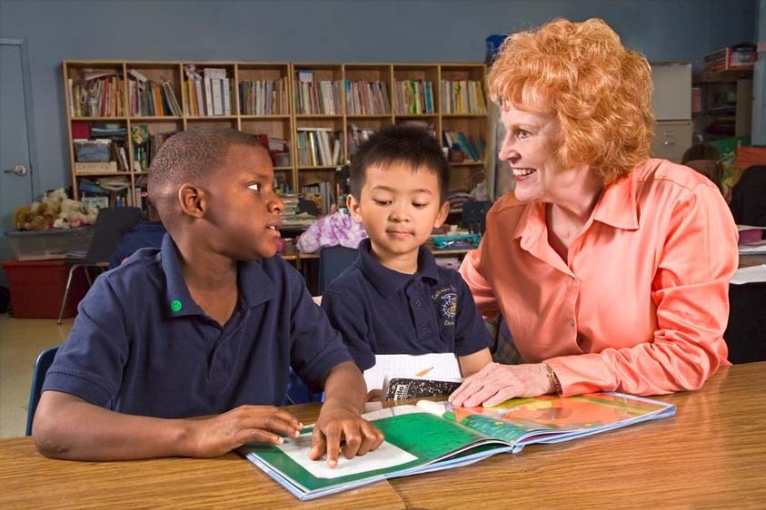 Caucasian female teacher with red hair shows excitement about black male student's progress with reading while Vietnamese 7 boy looks on Teacher shows approval of black boy reading book in second grade