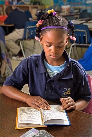 One black female second grader with braids in her hair and wearing school uniformreads a book at desk in classroom in New Orleans. African American girl 7 reads book in class at desk