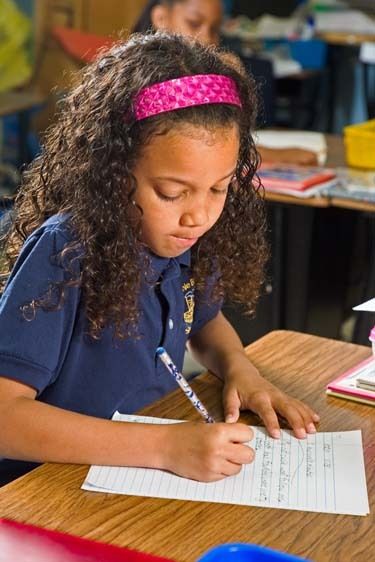 Seven year old mixed black female in uniform writes on lined paper at desk in classroom in New Orleans Mixed black second grade girl writes at desk