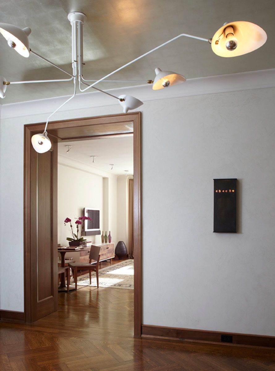 This apartment renovation features rich nut-wood veneers, walnut for the doors and pecan for the kitchen cabinetry, complemented with outsized patterned wallpaper in the entry vestibule and den.  The gallery is furnished with a Serge Mouille chandelier casting light on the silver leafed ceiling, a bronze wall-mounted BDDW vacuum tube clock, and a shagreen-fronted Roman Thomas console.Construction was completed in 2006, and Belsey & Mahla Architects continues to provide design services to the owners. West 86th Street