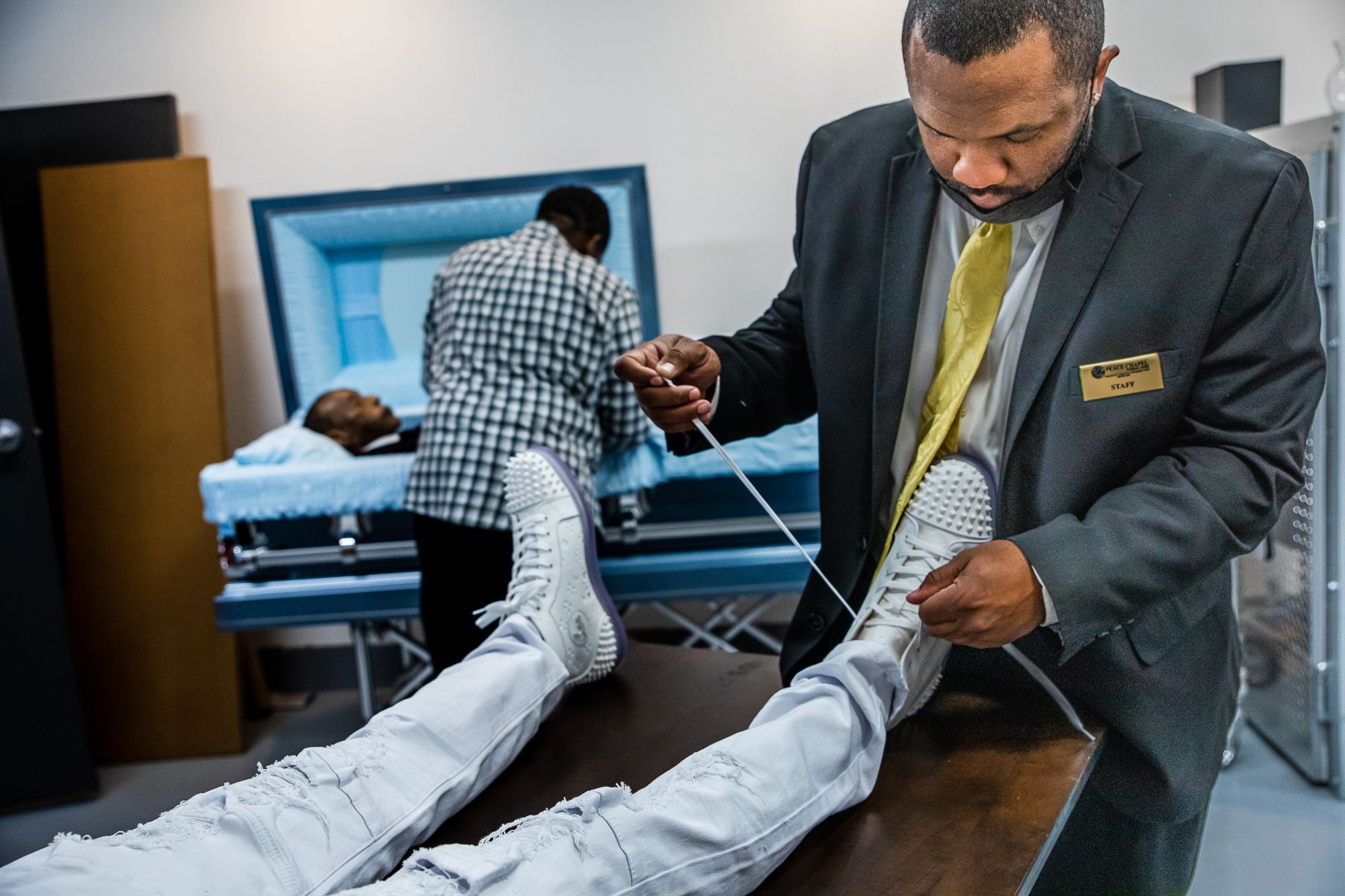 As funeral director Ron Thornson works behind him, Reginald Burgan ties a deceased man's shoelaces at Peace Chapel Funeral Home in Phoenix on March 12, 2021. Burgan, who is the funeral home’s office manager, was hired in August to run the office so Thornson could focus on his embalming work. 2021_0423_FinalProject_Mariani-014.jpg