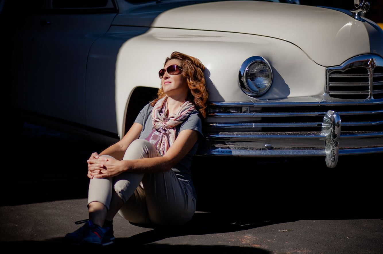 The woman is sitting by the old car