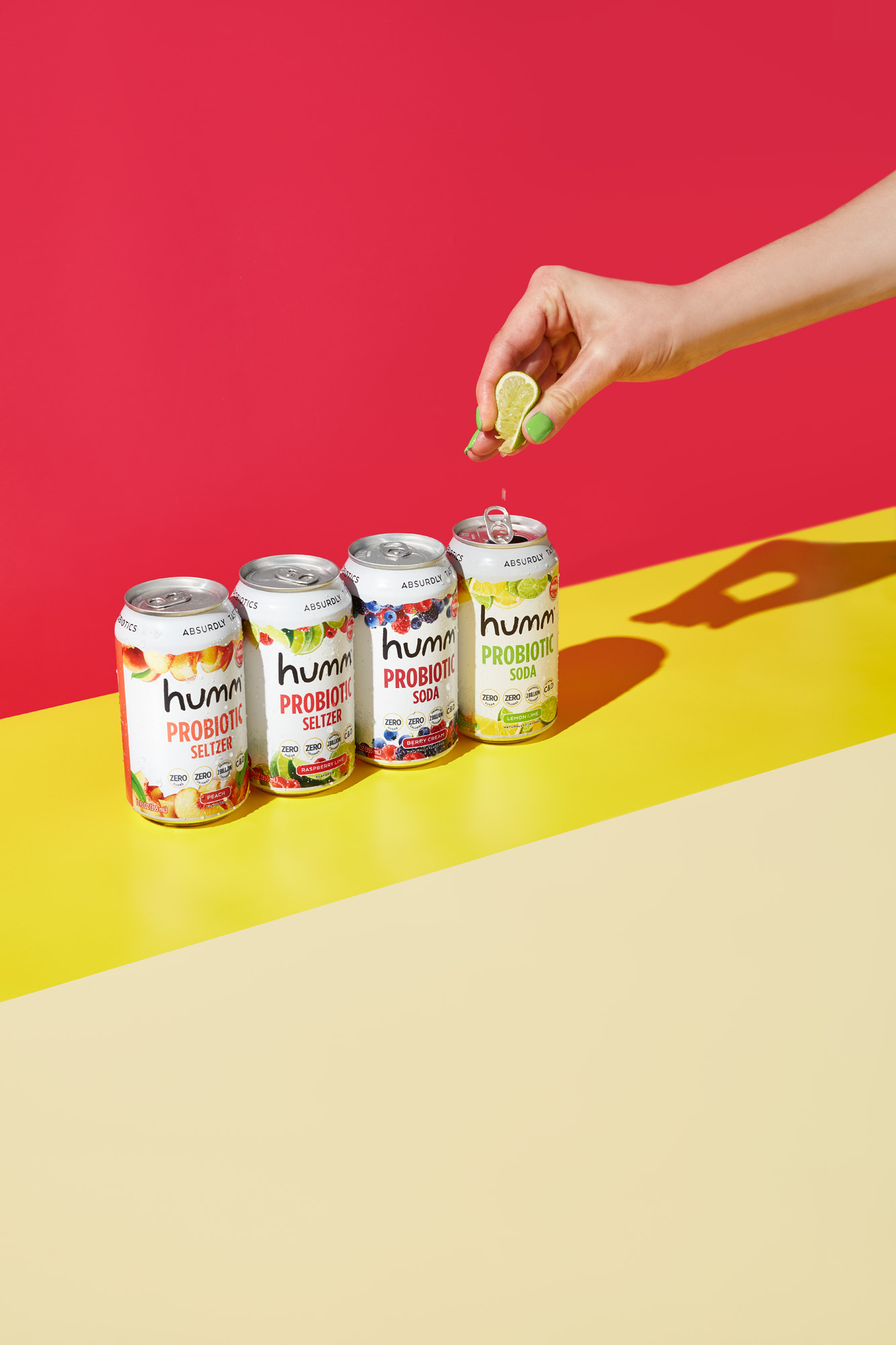 A variety of Humm Kombucha flavors lined up on a vibrant yellow and pink backdrop with a hand squirting lime into the probiotic drink Probiotic Kombucha