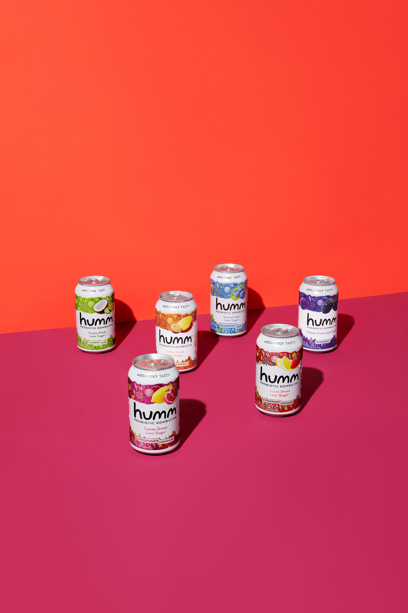A variety of Humm Kombucha flavors lined up on a vibrant yellow and pink backdrop with a hand squirting lime into the probiotic drink Probiotics Kombucha Drink