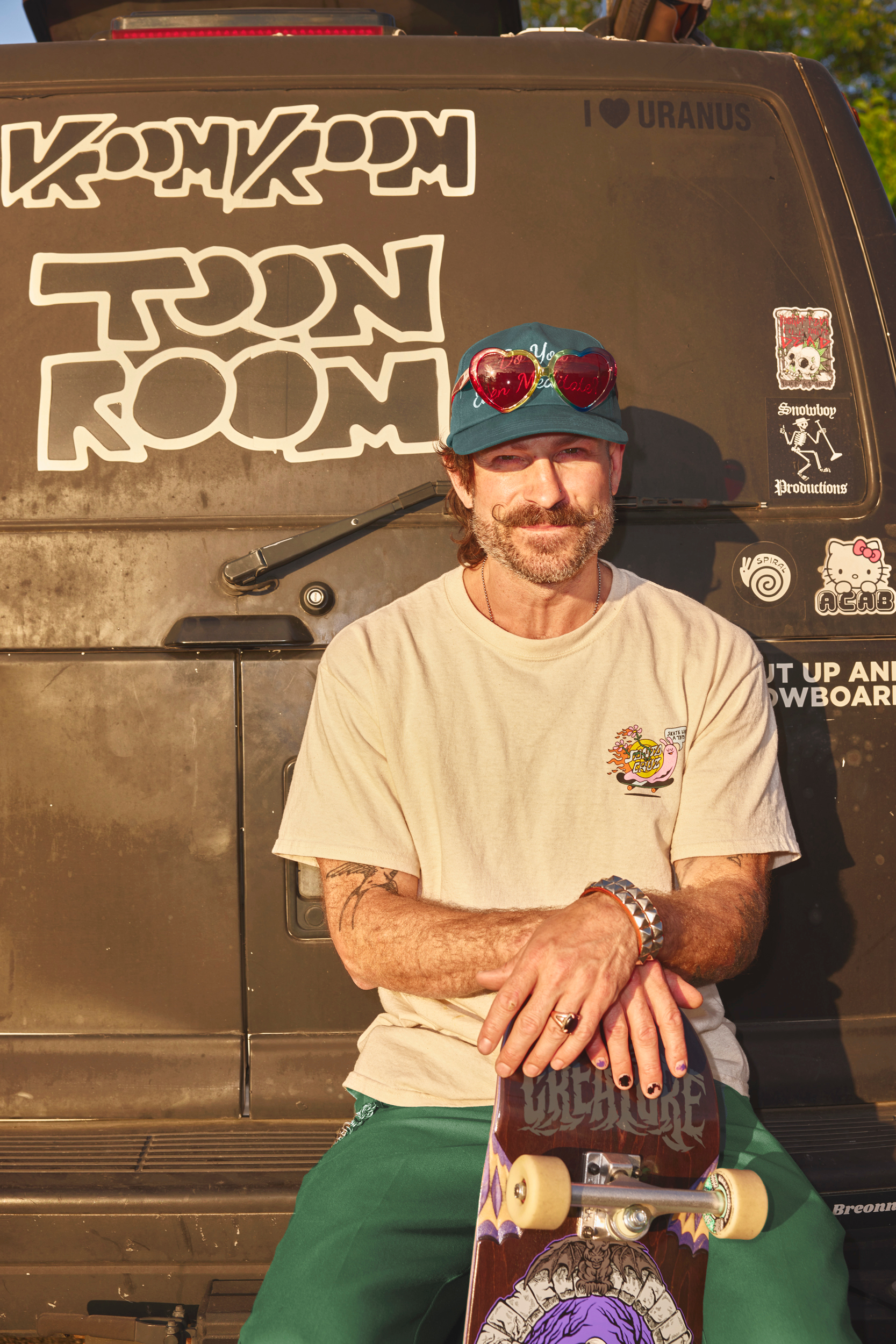 hipster man poses against a van with skateboard and heart sunglasses