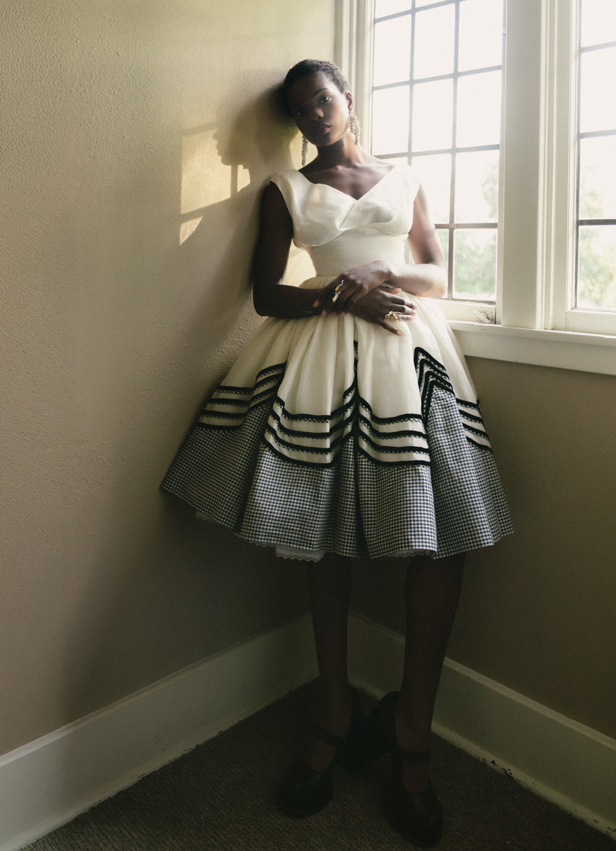 black model with buzz cut in black and white 50s style dress leans against window 