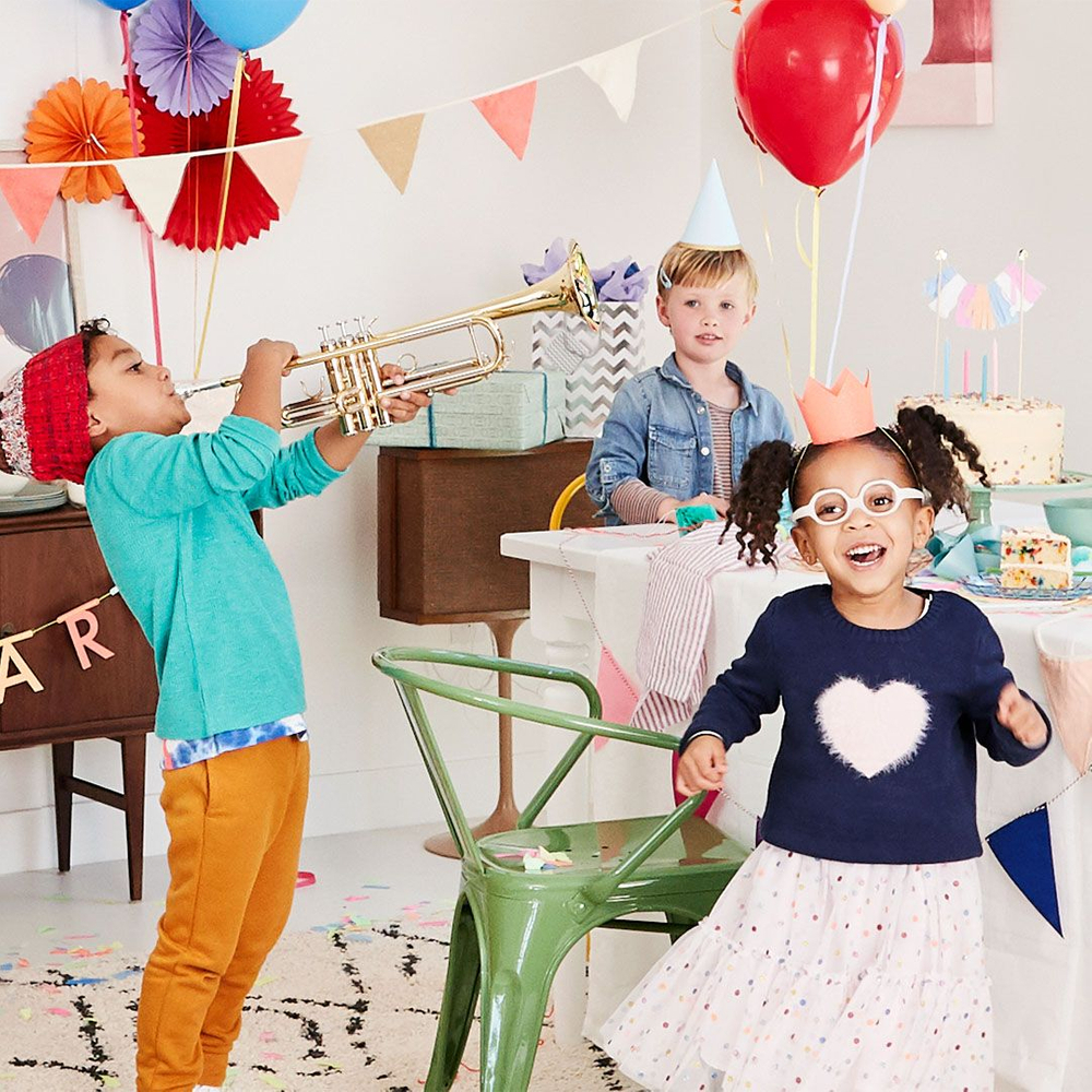 little boy plays a trumpet, little girl in party hat dances, boy in party hat waits to blow out candles at birthday party three kids celebrating around a table at a birthday party