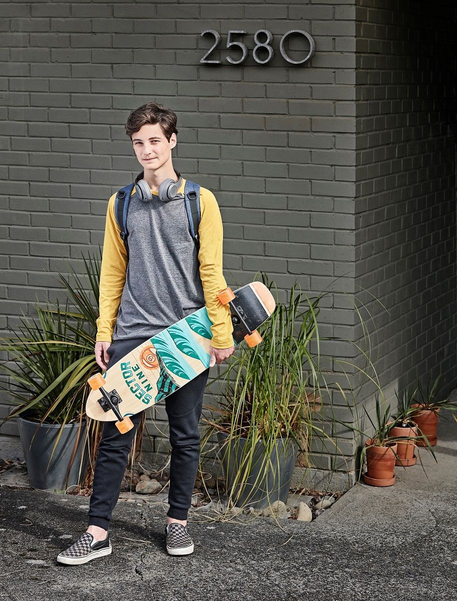 teenage boy wearing a backpack and head phones hold his skateboard outside of a house. photo retouching by jon birdseye teenage boy holds skate board outside of house