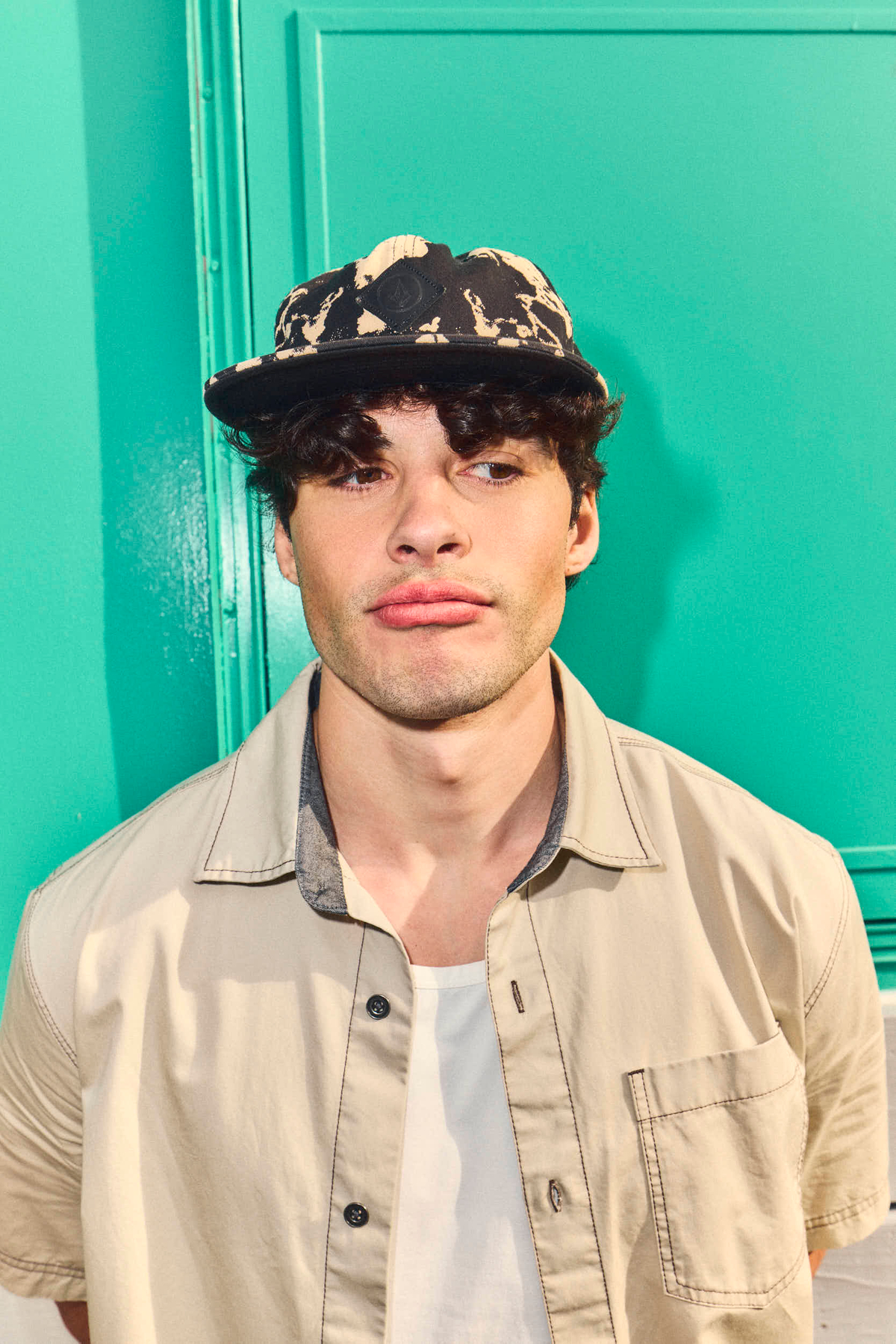 young hispanic gen z man with curly hair leans against seafoam green wall