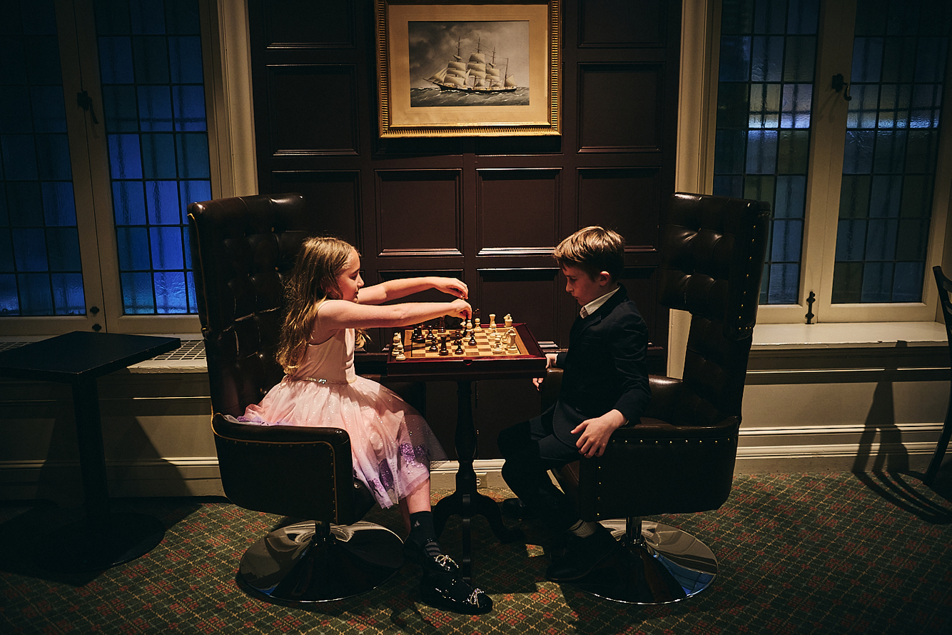little girl in pink tutu and boy in suit play chess in ornate study  young siblings play chess
