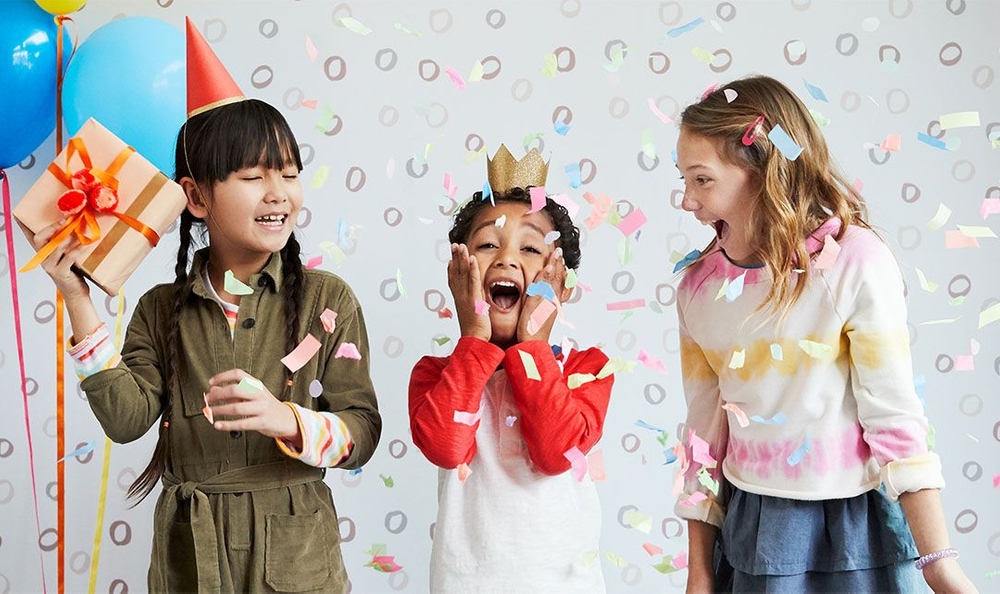 young boy standing between two young girls laughing as confetti falls at a birthday party three kids in birthday hats laughing at a birthday party