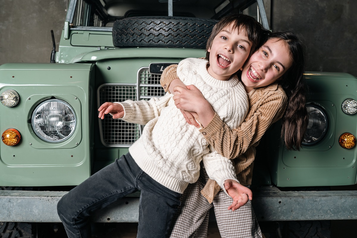 young siblings laugh and giggle as they play in front of a vintage jeep 