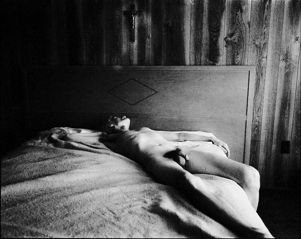 Black-and-white photograph of a nude figure reclining on a motel bed in a wood-paneled room, eyes turned toward a cross above the headboard, exploring themes of faith, vulnerability, and desire from Micha Warren’s series God Is Never Far Away. Nude figure in wood-paneled motel room gazing at cross – “God Is Never Far Away”