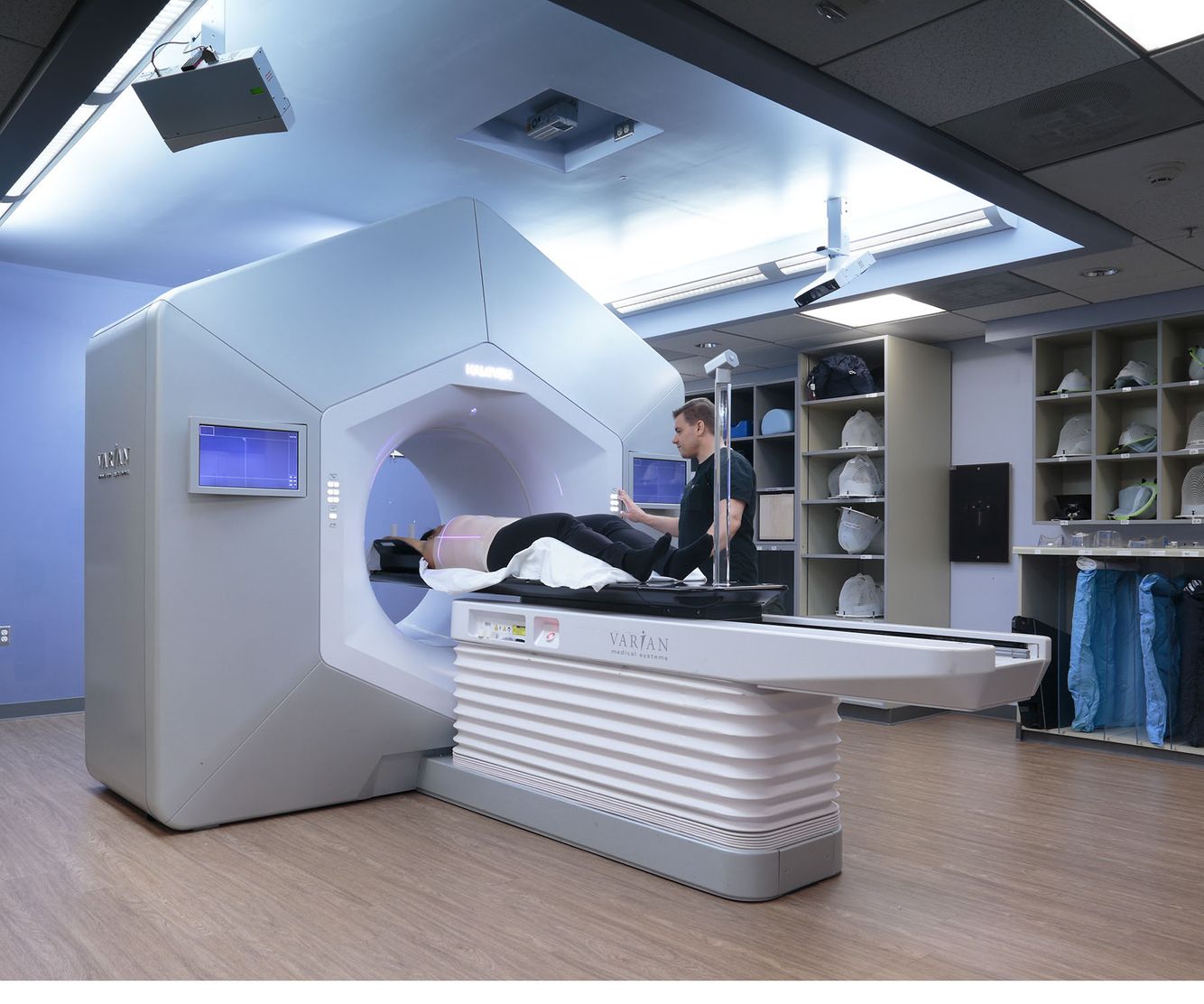 A clean, technically precise commercial photograph of an advanced medical treatment system in a modern clinical suite. A clinician is demonstrating a patient setup on a Varian linear accelerator. Life Sciences & Medical Technology Clinical Suite