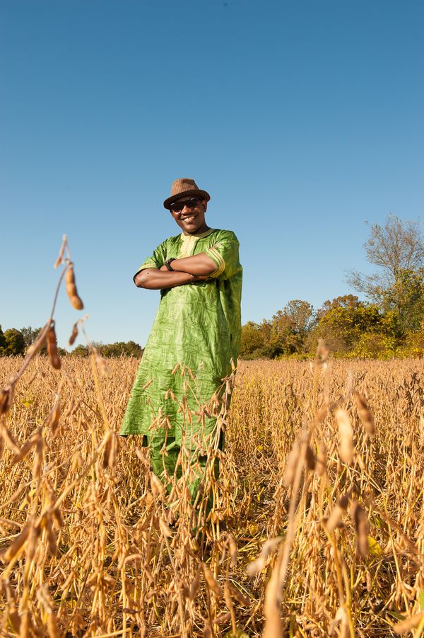 Mamdou Kelly singer and guitar player from Mail stands in a field of golden dried alphpha and wearing a long green African dashiki, brown fedora, and sun glasses Mamadou Kelly Musician from Mali