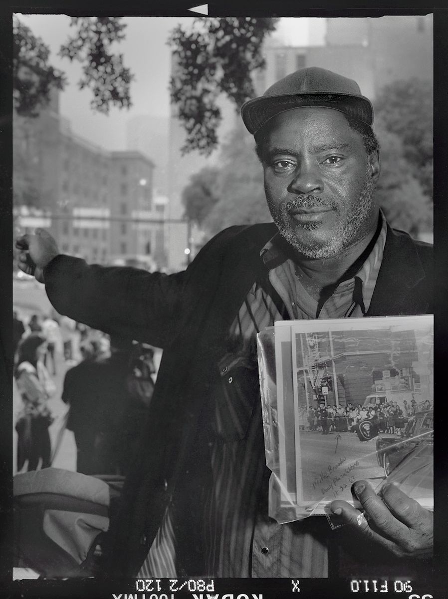 MIKE BROWNLOW, WAS A 13-YEAR OLD AT THE MOTORCADE PARADE; HERE, POINTING TO WHERE HE STOOD AND SAW GUNFIRE COME FROM BEHIND PICKET FENCE, ON THE GRASSY KNOLL