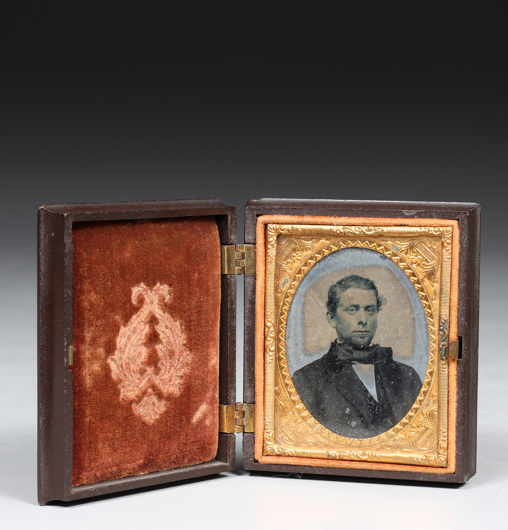An antique daguerreotype portrait of a man in a hinged leather case with red velvet lining and ornate gold decorative mat, photographed against a dark gradient background. Antique collectible auction photography by Victor Koroma for Auctions. 9th Century Daguerreotype Portrait Gilt Case — I.M. Chait Auction Photography