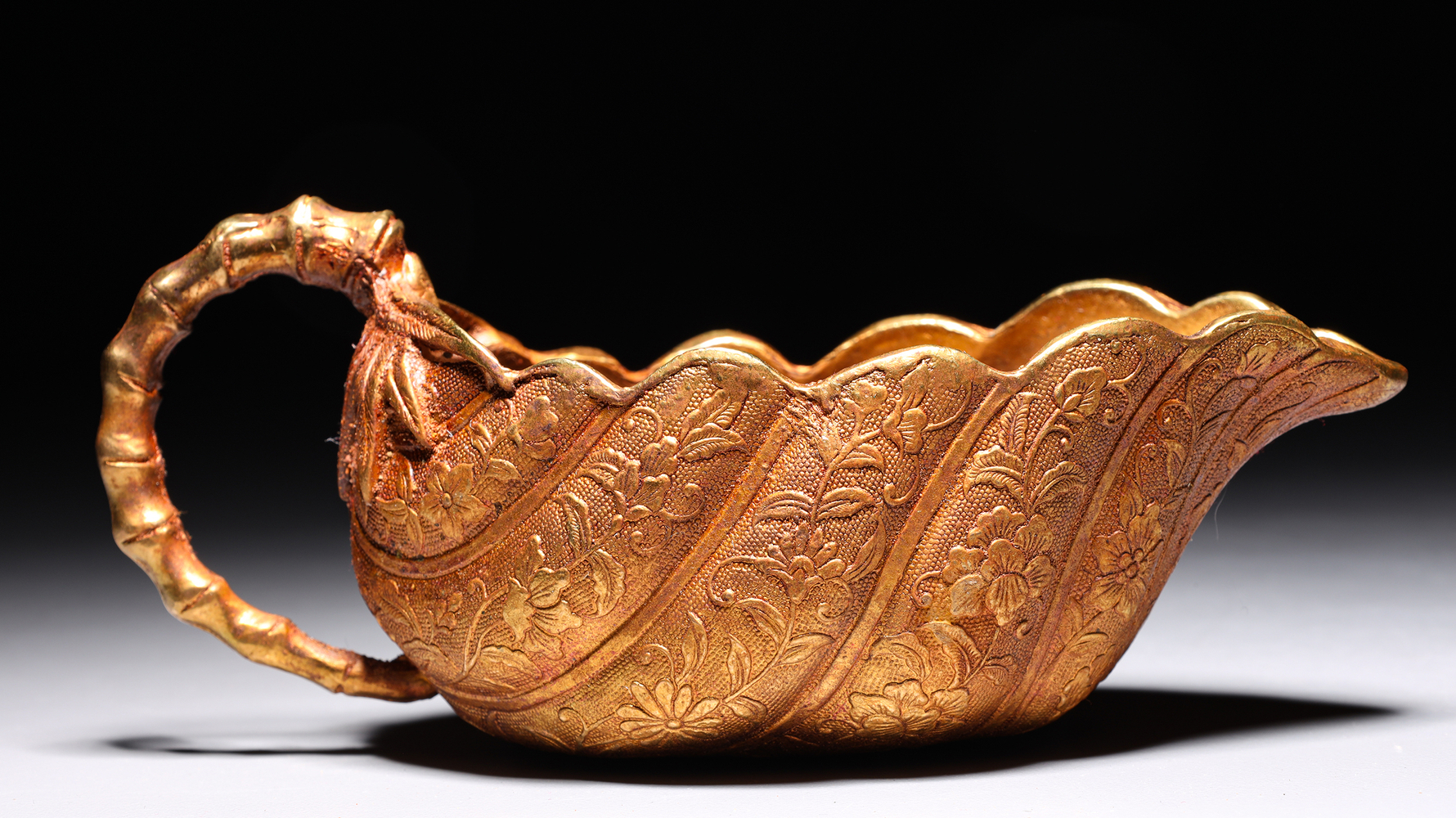 Auction photography detail of a Qing Dynasty gilded bronze cup with repoussé floral decoration, spiral ribbing, scalloped rim and bamboo-form handle against black background. Photographed for I.M. Chait Gallery and Auctioneers and featured on LiveAuctioneers by Los Angeles commercial photographer Victor Koroma. Qing Dynasty Gilded Bronze Cup Detail — I.M. Chait LiveAuctioneers Victor Koroma