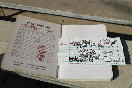 Fine art photograph of an open wide-ruled composition notebook featuring a hand-drawn Basquiat-inspired skull with the text "Most Young Kings Get Their Guns Off Head," crossed-out words, a crown, and the word "Revenge," with art class homework reading "Write a paragraph about how the art world viewed Basquiat," photographed on a concrete surface. From the My Middle School Notebook series by Los Angeles photographer Victor Koroma. Basquiat Art Class — MY MIDDLE SCHOOL NOTEBOOK | Fine Art Photography | Victor Koroma