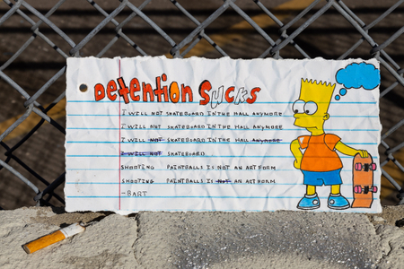 Fine art photograph of a torn notebook page reading "Detention Sucks" with a hand-drawn Bart Simpson holding a skateboard and detention lines including "I will not skateboard in the hall anymore" and "Shooting paintballs is not an art form," propped against a chain-link fence with a cigarette butt on concrete below. From the My Middle School Notebook series by Los Angeles photographer Victor Koroma.
Bart Simpson Detention Sucks — MY MIDDLE SCHOOL NOTEBOOK | Fine Art Photography | Victor Koroma