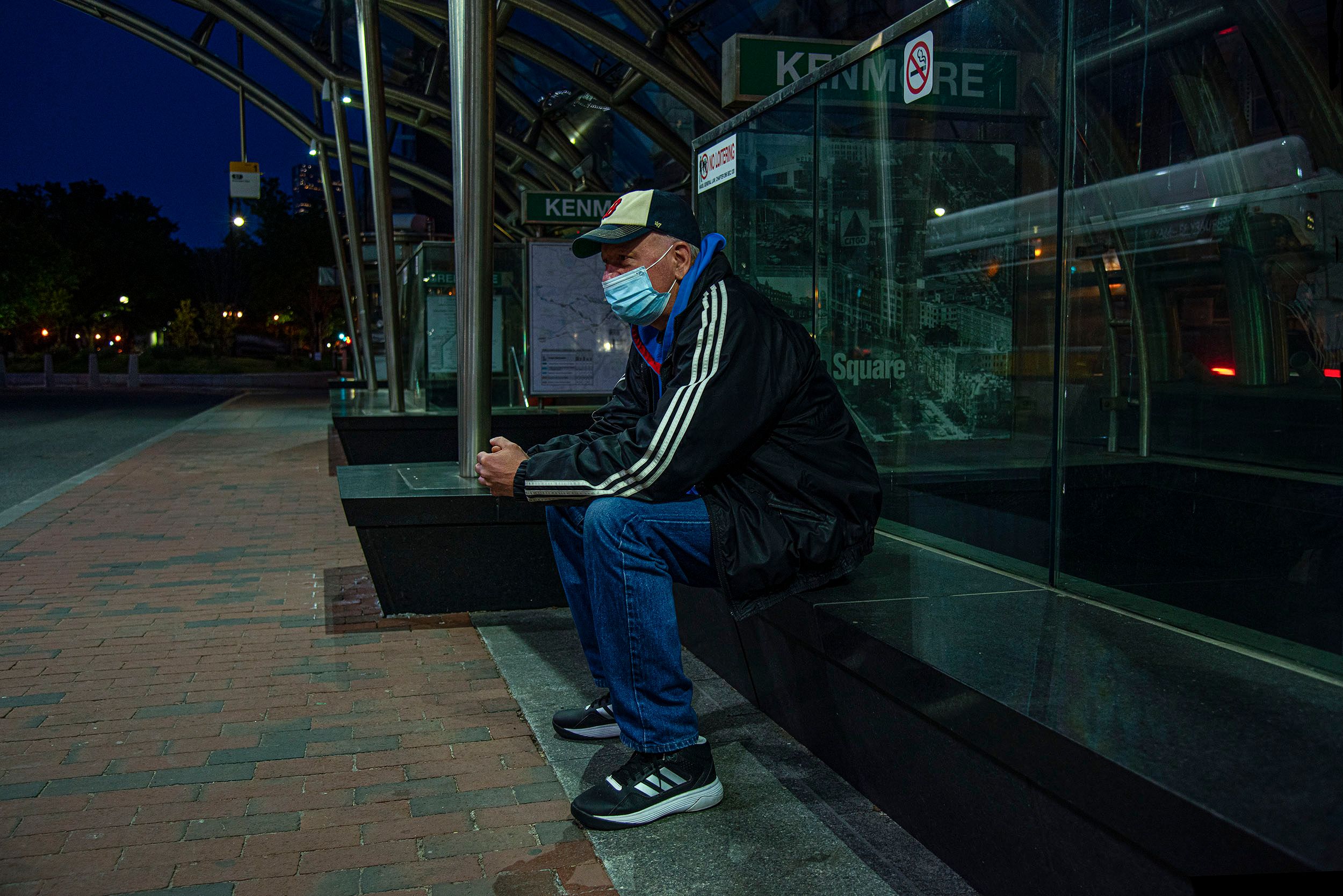 Photograph of a man at night wearing a facemask sitting on a bench and waiting for a bus at the Kenmore Bus station during coronavirus pandemic. Boston, MA Waiting for the bus