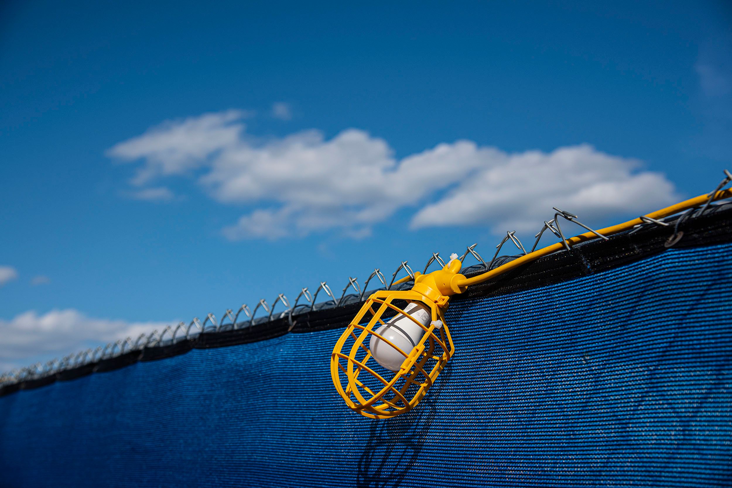 Horizontal color photograph of a light bulb encased in a yellow plastic housing  and hanging on a fence with blue sky and clouds in the background. Lightbulb