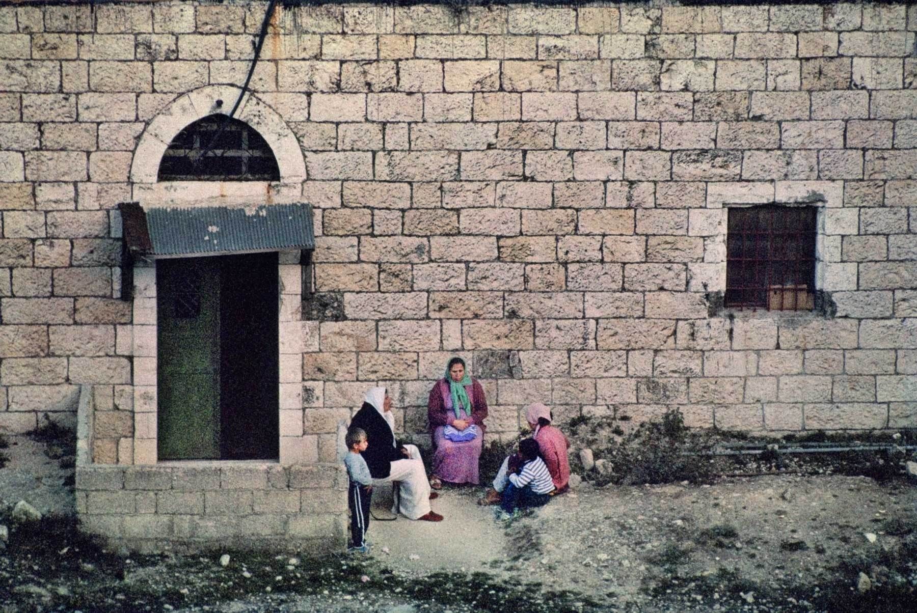 Palestinian women and children sit and chat against a background of age-old Jerusalem stone, in a view looking down from the old city's rocky slopes. Conversation, the Old City