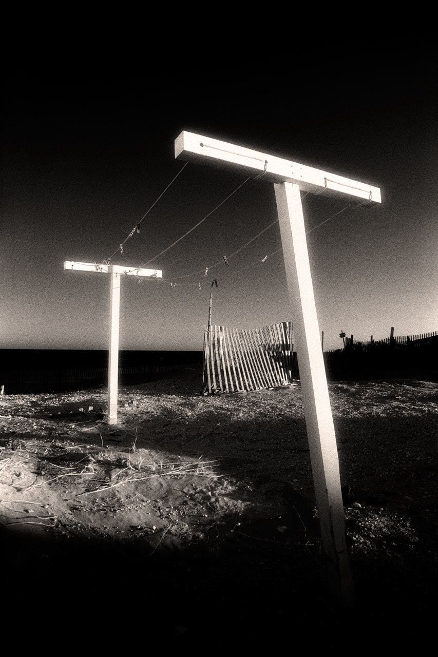 A clothesline near the beach on Cape Cod, Massachusetts, photographed by very low winter sun. The light in this image reminds me of a movie set, an effect enhanced by the way the horizon's glow (caused by my red filter's graduated darkening of the blue sky) silhouettes erosion-control fencing at the center right of the image. The black band behind the far support is the sea itself, also darkened by the filter with help in printing. 1tbars_final