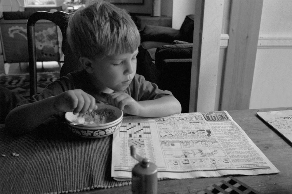 Boy  eating cereal with hand reading comic Sam Roos Cape Elizabeth Maine B&W street photography 1994 nineteen  nineties
