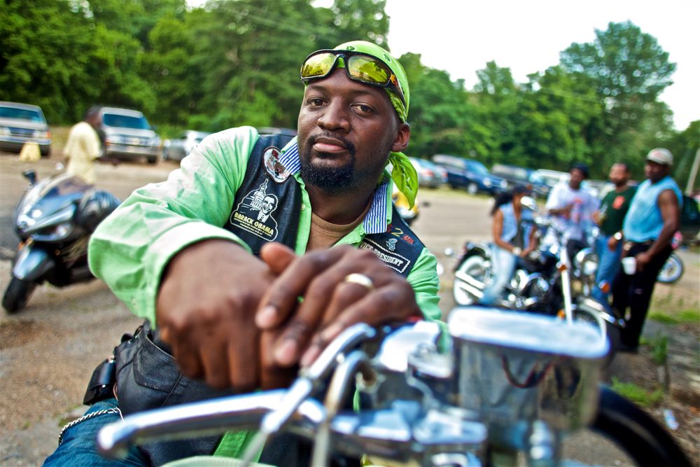 Environmental portrait of a motorcycle rider dressed in green with a black vest on looking into the camera over his handle bars at a music festival. Other riders mingle in the background. Photos of various people & locations on and around the delta on Highway 61, The Blues Highway that runs north from Memphis, Tennessee to New Orleans, Louisiana. Shot for a number of projects such as the documentary We Juke Up In Here, the web series Moonshine & Mojo Hands, a passion project to document the beautiful Mississippi lifestyle. As an on location photographer, lifestyle photographer & location commercial photographer, this was one the best long term projects ever that captures southern Americana! Motorcycle Rider in Green
