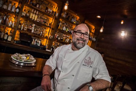 Portrait of Robert St. John smiling looking off camera at the bar at his Purple Parrot restaurant in Hattiesburg , MS. He is a CHEF, AUTHOR, RESTAURANTEUR, & WORLD-CLASS EATER, Mississippi. Shot for the Mississippi Tourism campaign for advertising, branding, collateral and broadcast motion assets. Environmental portrait, and we are a industrial photographer, on location photographer & location commercial photographer. Shallow depth of field and selective focus. VisitMS Portrait of Robert St. John CHEF, AUTHOR, RESTAURANTEUR, & WORLD-CLASS EATER