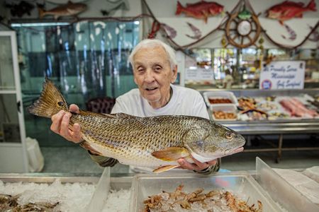 A man, purveyor in a fish market holding one of his fish for sale, with a smiling confident pleasant expression. Shot for the Mississippi Tourism campaign for advertising, branding, collateral and broadcast motion assets. Environmental portrait, and we are a industrial photographer, on location photographer & location commercial photographer. Shallow depth of field and selective focus. VisitMS Man Holding Fish at a Fish MArket