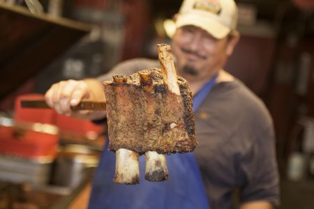 A photo of a man holding a slab of ribs to the camera and smiling. The man is smiling with a pleasant confident expression. Shot for the Mississippi Tourism campaign for advertising, branding, collateral and broadcast motion assets. Environmental portrait, and we are a industrial photographer, on location photographer & location commercial photographer. Shallow depth of field and selective focus. Man Holding Ribs Toward the Camera