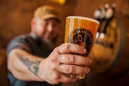A photo of a man holding a freshly poured draught beer at the Crooked Letter Brewery on Mississippi's Gulf Coast. He is holding it toward the camera and smiling and the beer is the emphasis of the shot and focus. Shot for the Mississippi Tourism campaign for advertising, branding, collateral and broadcast motion assets. Environmental portrait, and we are a industrial photographer, on location photographer & location commercial photographer. Shallow depth of field and selective focus. Mn Holding a Glass of Beer toward the Camera